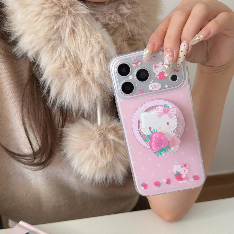 May include: A pink phone case featuring a Hello Kitty design with a strawberry. The case has a clear border and a pop-up grip with the Hello Kitty character. The background is dotted with small pink circles and strawberry illustrations.