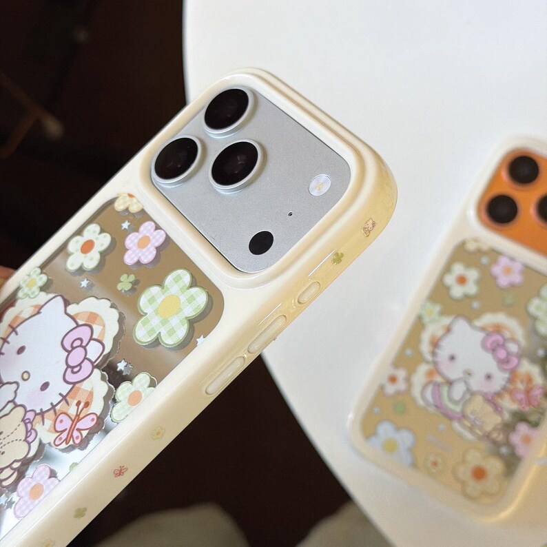 May include: Cream-colored phone case featuring a Hello Kitty design with flowers and butterflies. The case has a clear section with the character and floral elements. The phone case is shown with a second case with an orange top.