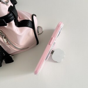May include: A pink phone case with a clear back and a white pop-up grip is shown. The phone case is next to a small pink and black bag with silver hardware. The bag has a zipper and a black strap.