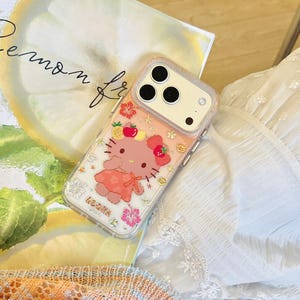 May include: A clear phone case featuring a pink cartoon character with a red bow and dress, surrounded by fruit and flower designs. The case has a pink border and is on a book with the words "Lemon fr" visible.