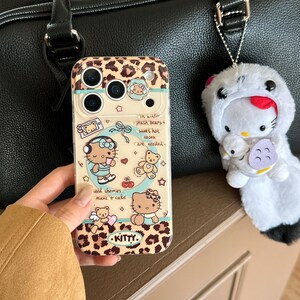 May include: A clear phone case with a leopard print design and illustrations of Hello Kitty characters. The case has text that reads "In winter, plash bears + sweet hot cocoa are needed." A Hello Kitty plushie keychain is attached.
