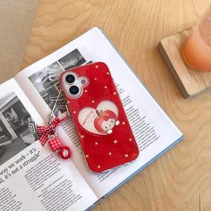 Puede incluir: Funda de tel&eacute;fono roja con dise&ntilde;o de Hello Kitty. La funda presenta un recorte en forma de coraz&oacute;n con un gr&aacute;fico de manzana y Hello Kitty, rodeado de estrellas doradas. Incluye un charm rojo y blanco.