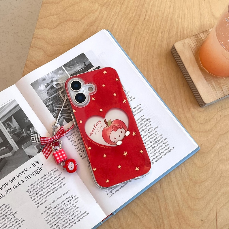Puede incluir: Funda de tel&eacute;fono roja con dise&ntilde;o de Hello Kitty. La funda presenta un recorte en forma de coraz&oacute;n con un gr&aacute;fico de manzana y Hello Kitty, rodeado de estrellas doradas. Incluye un charm rojo y blanco.