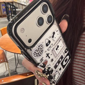Op de afbeelding: Een zwarte en doorzichtige telefoonhoes met een decoratief patroon met harten, strikken en andere kleine iconen. De hoes heeft een zwarte rand en een zilveren cameragedeelte.