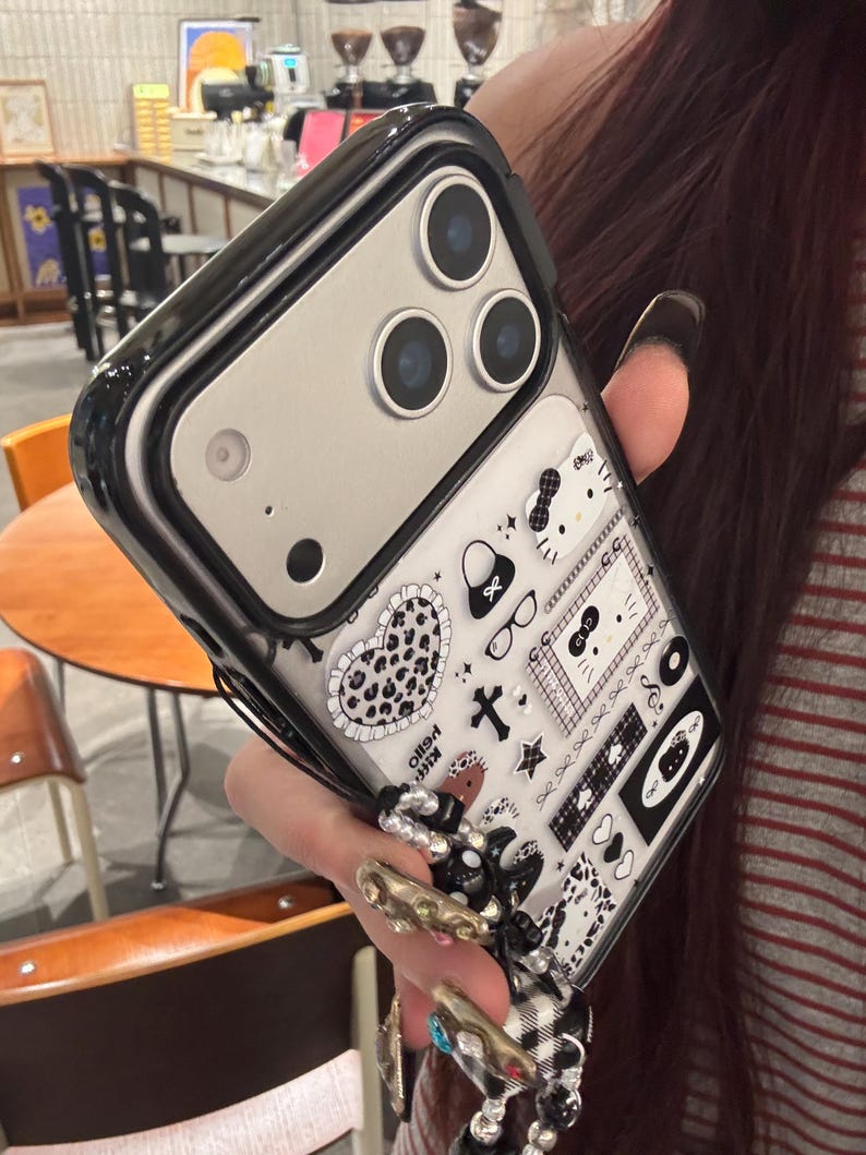 Op de afbeelding: Een zwarte en doorzichtige telefoonhoes met een decoratief patroon met harten, strikken en andere kleine iconen. De hoes heeft een zwarte rand en een zilveren cameragedeelte.