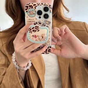 May include: Clear phone case with a leopard print design and a Hello Kitty theme. The case features a pop-up grip with a Hello Kitty design and a collection of teddy bears. The case is held by a person wearing a bracelet.