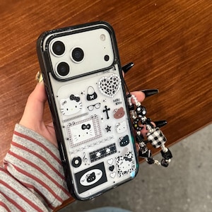 Op de afbeelding: Zwarte en transparante telefoonhoes met een Hello Kitty-ontwerp. De hoes heeft verschillende zwart-witte afbeeldingen, waaronder harten, strikken en het Hello Kitty-personage. Een kralenbedel is aan de zijkant bevestigd.