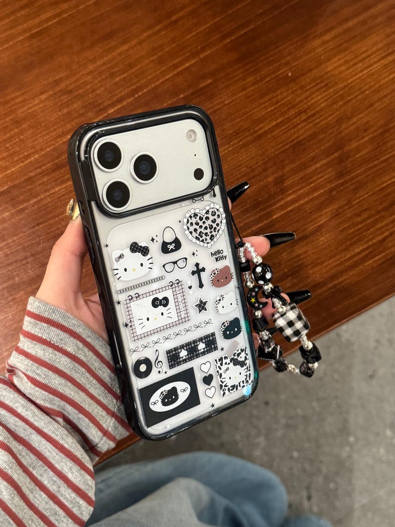 Op de afbeelding: Zwarte en transparante telefoonhoes met een Hello Kitty-ontwerp. De hoes heeft verschillende zwart-witte afbeeldingen, waaronder harten, strikken en het Hello Kitty-personage. Een kralenbedel is aan de zijkant bevestigd.