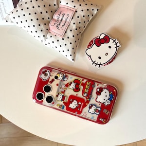 May include: A red and clear phone case featuring Hello Kitty designs, a white and black polka dot pillow, and a Hello Kitty phone grip. The phone case has a red border and a clear center with multiple Hello Kitty graphics.