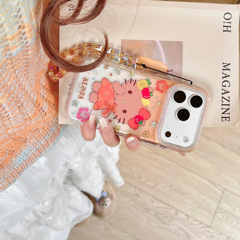 May include: Clear phone case with a pink border featuring a cartoon character, strawberries, and the word "Aloha". The case has a gold chain strap and is held by a person.