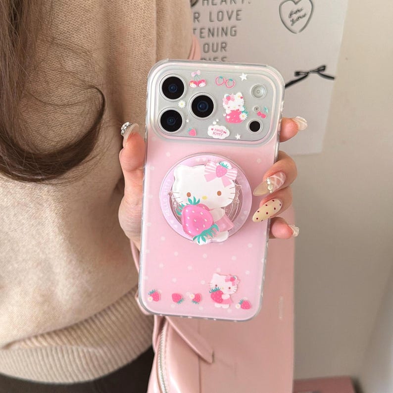 May include: Pink phone case with a Hello Kitty design. The case features a pop-up grip with Hello Kitty holding a strawberry, and is decorated with small Hello Kitty and strawberry graphics. The case has a clear border and is held in a person's hand.
