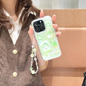 May include: Clear phone case with a green apple design and a beaded wrist strap. The case features cartoon illustrations of apples and other green elements. The wrist strap has green and clear beads and a star charm.