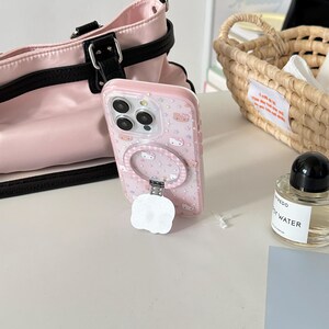 May include: A pink phone case with a ring holder, decorated with a repeating pattern of cartoon characters and stars. The case is shown with a pink handbag and a small perfume bottle.