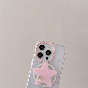 K&ouml;nnte beinhalten: Transparente Handyh&uuml;lle mit einem rosa sternf&ouml;rmigen Handyhalter. Die H&uuml;lle hat einen rosa Rand um die Kamera und einen Ringhalter mit goldenem Schleifendetail. Die H&uuml;lle hat ein Muster aus kleinen Sternen.