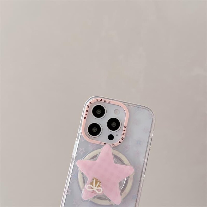 K&ouml;nnte beinhalten: Transparente Handyh&uuml;lle mit einem rosa sternf&ouml;rmigen Handyhalter. Die H&uuml;lle hat einen rosa Rand um die Kamera und einen Ringhalter mit goldenem Schleifendetail. Die H&uuml;lle hat ein Muster aus kleinen Sternen.