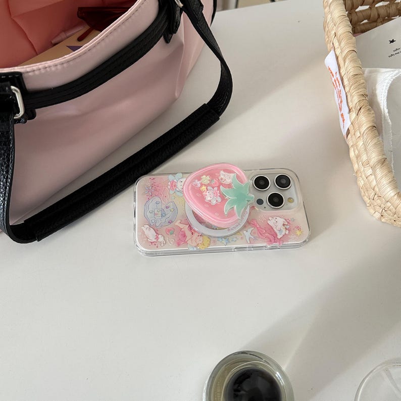 May include: A clear phone case with a pink strawberry-shaped pop socket. The case features a colorful design with cartoon characters. A pink purse with a black strap is in the background.