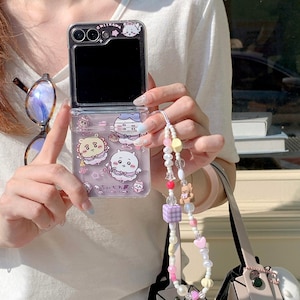 May include: A clear foldable smartphone case featuring Chikawa character graphics in purple and white, with the text CHIIKAWA printed at the top. Attached is a beaded phone charm strap with white pearls, pink hearts, a small bear, and a purple cube.