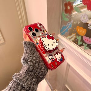May include: A red and clear phone case featuring Hello Kitty designs. The case has a pop-up grip with a Hello Kitty face and a red bow. The case is decorated with various Hello Kitty characters and accessories.