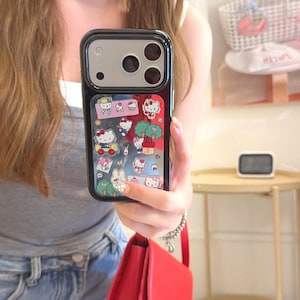 May include: Black phone case with a colorful Hello Kitty design. The case features a clear window with various Hello Kitty illustrations, including the character in a car, near trees, and with other characters. The case has a black border and a camera cutout.