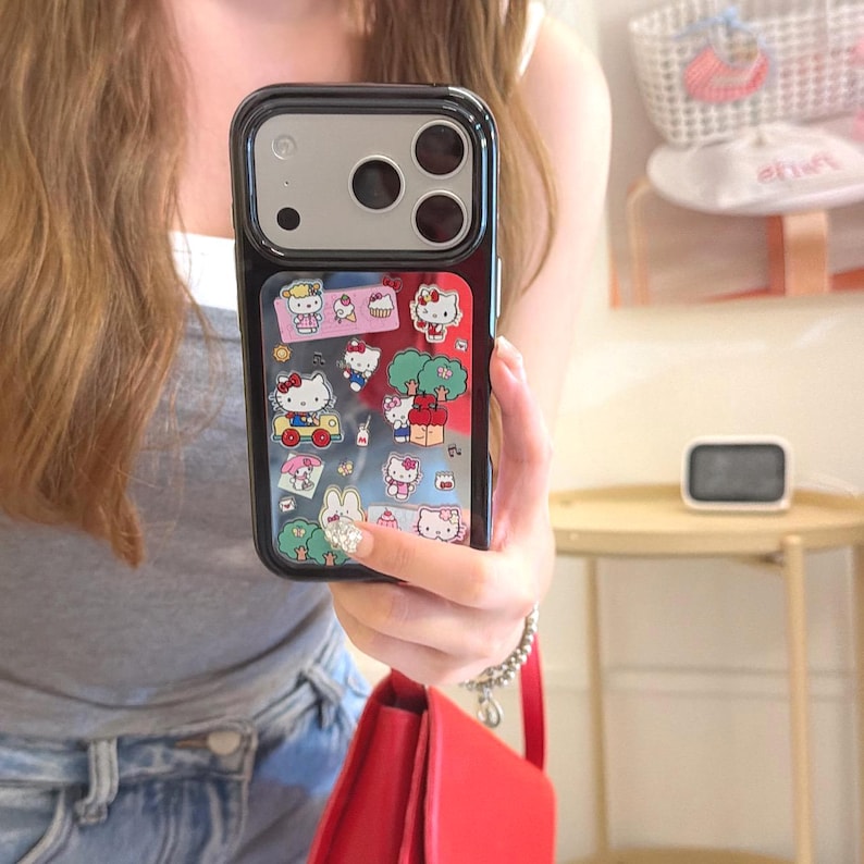 May include: Black phone case with a colorful Hello Kitty design. The case features a clear window with various Hello Kitty illustrations, including the character in a car, near trees, and with other characters. The case has a black border and a camera cutout.