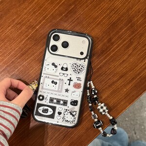 Op de afbeelding: Transparante telefoonhoes met zwarte rand, versierd met een Hello Kitty-ontwerp en diverse iconen. De hoes heeft een zwart-witte kralen polsband.