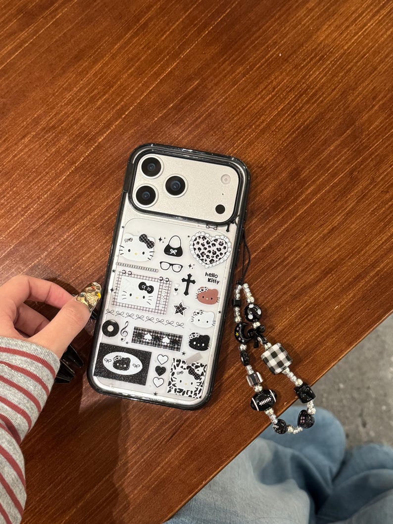Op de afbeelding: Transparante telefoonhoes met zwarte rand, versierd met een Hello Kitty-ontwerp en diverse iconen. De hoes heeft een zwart-witte kralen polsband.