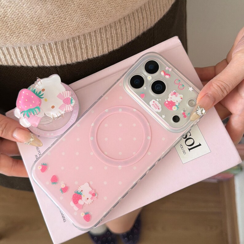 May include: Pink iPhone case with a Hello Kitty design, featuring a strawberry and bow accent. The case has a clear, protective cover with a ring for MagSafe charging. A matching phone grip with a Hello Kitty and strawberry design is also visible.