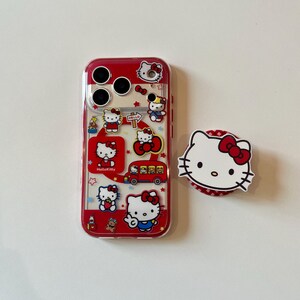 May include: A clear phone case with a red border and Hello Kitty graphics. The case features multiple Hello Kitty images, stars, and a red bus. A matching white and red Hello Kitty phone grip is also present.