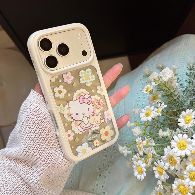 May include: A cream-colored phone case featuring a clear window with a floral pattern and a cartoon character. The case has a protective bumper around the camera lenses. The design includes a cartoon cat with a bow.