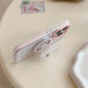 K&ouml;nnte beinhalten: Eine transparente Handyh&uuml;lle mit rosa Rand und passendem Pop-up-Griff. Die H&uuml;lle hat einen rosa Ring um die Kamera und einen transparenten, bl&uuml;tenf&ouml;rmigen Griff. Die Handyh&uuml;lle liegt auf einer hellen Oberfl&auml;che.