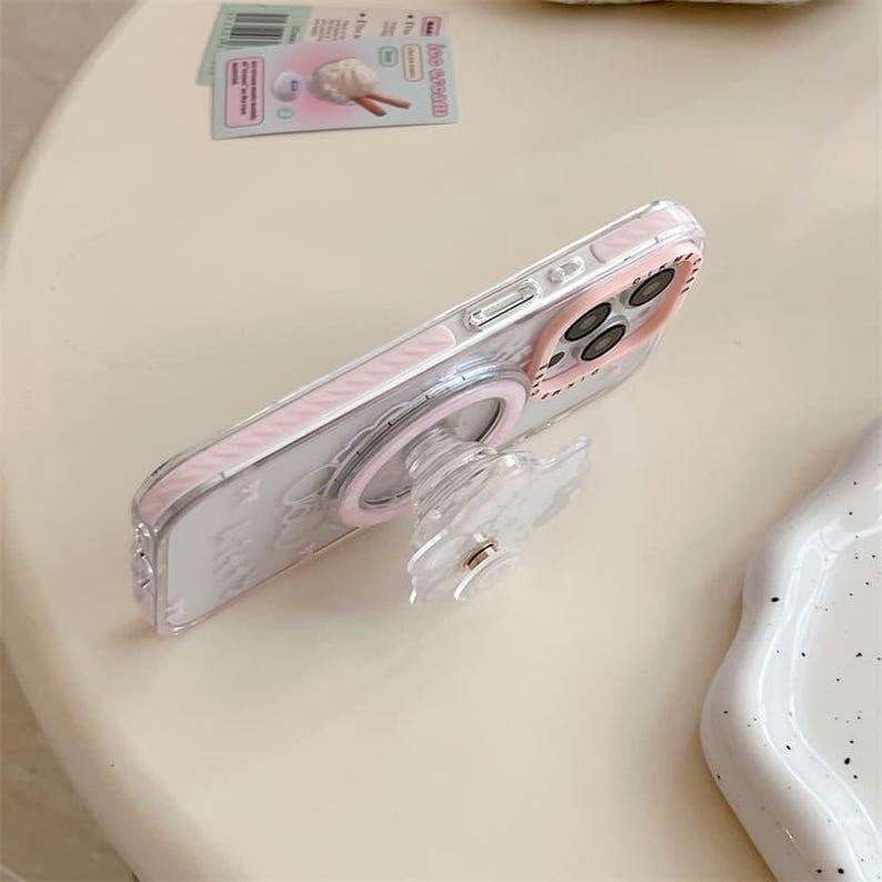 K&ouml;nnte beinhalten: Eine transparente Handyh&uuml;lle mit rosa Rand und passendem Pop-up-Griff. Die H&uuml;lle hat einen rosa Ring um die Kamera und einen transparenten, bl&uuml;tenf&ouml;rmigen Griff. Die Handyh&uuml;lle liegt auf einer hellen Oberfl&auml;che.