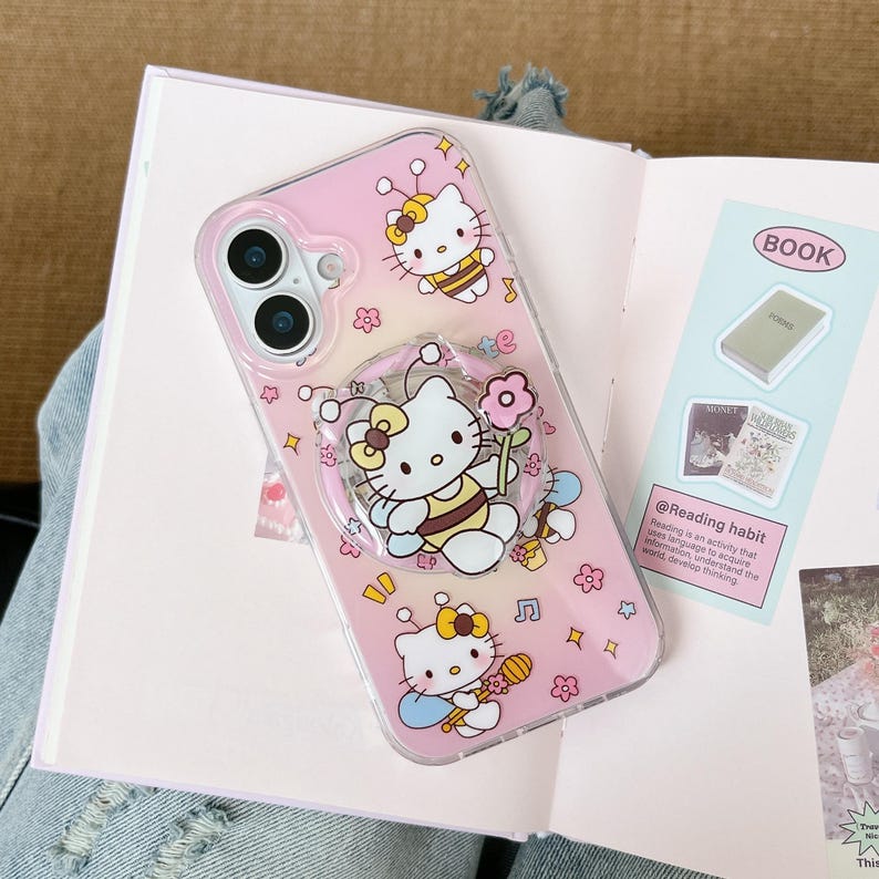 May include: A clear phone case with a pink gradient design featuring Hello Kitty characters dressed as bees. The case has a pop-up grip and is decorated with flowers, musical notes, and stars. The phone case is on top of an open book.