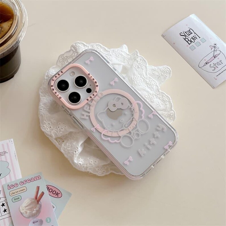 K&ouml;nnte beinhalten: Transparente Handyh&uuml;lle mit rosa Rahmen und Cartoon-Design. Die H&uuml;lle zeigt ein rosa Hello Kitty-Design mit Schleifen und dem Wort "Kitty". Die H&uuml;lle liegt auf einer wei&szlig;en Spitzendecke.