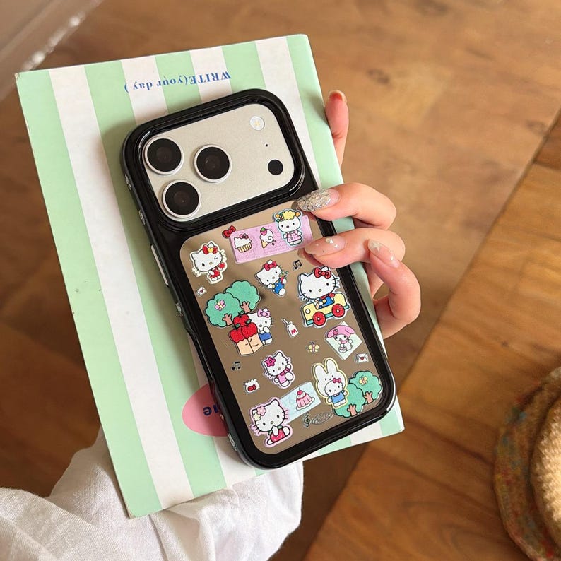 May include: Black phone case with a clear back featuring Hello Kitty stickers. The case has a black border and is held over a green and white striped notebook. The stickers include various Hello Kitty designs.