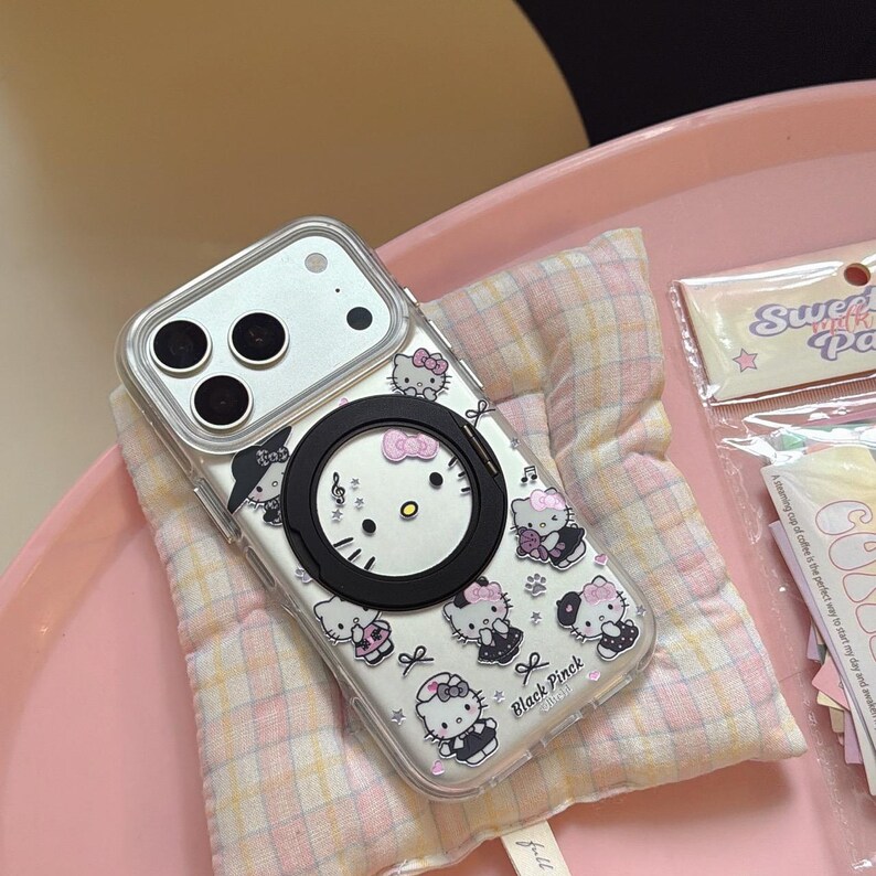 K&ouml;nnte beinhalten: Transparente Handyh&uuml;lle mit einem schwarzen Magnetring und einem Hello Kitty-Design. Die H&uuml;lle hat einen wei&szlig;en Hintergrund mit rosa Schleifen und Noten. Der Text "Black Pinck" ist sichtbar.