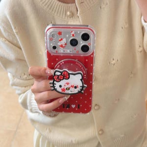 May include: Red phone case with a Hello Kitty design. The case features a pop-up grip with a Hello Kitty face and a red bow. The case has a red background with white stars and Hello Kitty images.