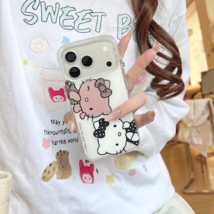 May include: Clear phone case featuring a pink and white cartoon character with a bow. The case has a clear border and is held in front of a white t-shirt with the words "Sweet B" and other graphics.
