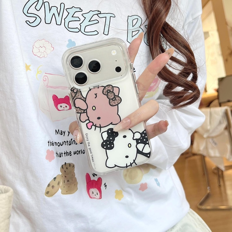 May include: Clear phone case featuring a pink and white cartoon character with a bow. The case has a clear border and is held in front of a white t-shirt with the words "Sweet B" and other graphics.