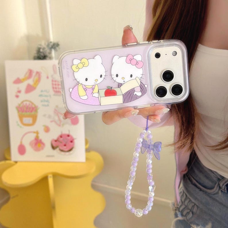 May include: A clear phone case featuring a purple background with two Hello Kitty characters. One Kitty wears a yellow bow and dress, the other a pink bow and striped outfit. A purple beaded wrist strap with a bow is attached.