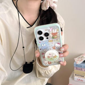May include: A clear phone case with a mint green border, featuring cartoon characters and a pop socket. The case has a variety of colourful illustrations, including bunnies, bears, and Hello Kitty. The pop socket has a glittery design.