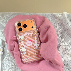 May include: Clear phone case with an orange camera section and a pink and white striped design. The case features Hello Kitty characters, bears, bows, and the word "HELLO" in a speech bubble. The phrase "SWEET HEART" is also visible.