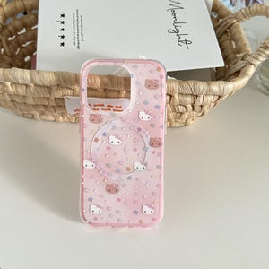 May include: A clear pink phone case with a pattern of Hello Kitty faces and stars. The case has a clear border and a circular design in the center. The case is on a white surface.
