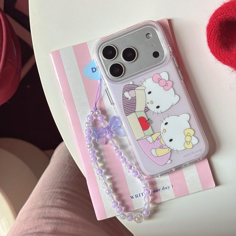 May include: A clear phone case with a purple Hello Kitty design. The case has a matching purple beaded wrist strap with a bow. The phone case is on a pink and white striped notebook.