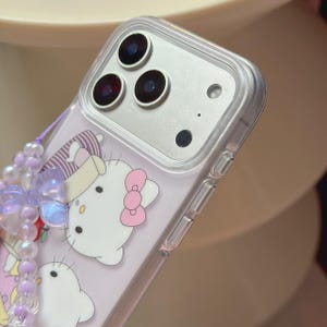May include: Clear phone case with a cartoon design featuring a white cat with a pink bow. The case has a beaded charm in shades of purple and white. The phone's camera lenses are visible.