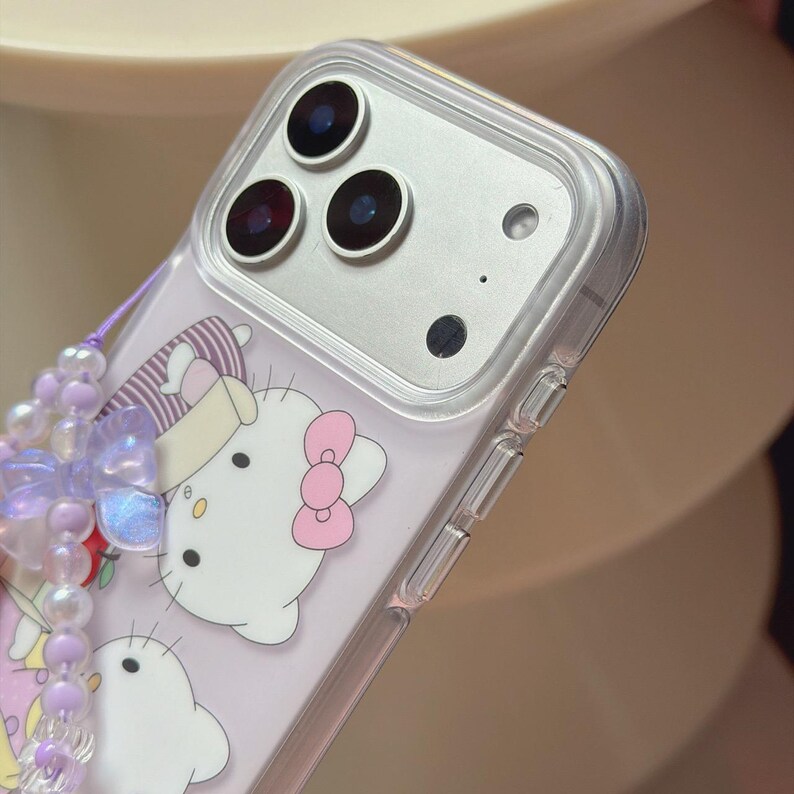 May include: Clear phone case with a cartoon design featuring a white cat with a pink bow. The case has a beaded charm in shades of purple and white. The phone's camera lenses are visible.