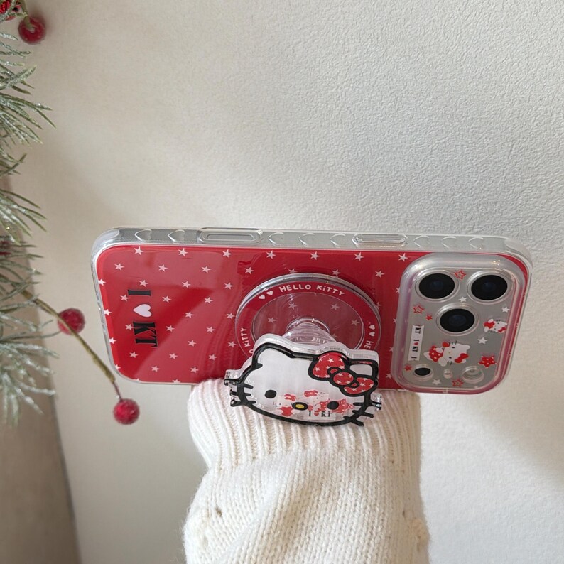 May include: Red phone case with a white star pattern and a Hello Kitty pop-up grip. The case has a clear border and the text "I KT" on the side. The pop-up grip features a Hello Kitty design with a red bow.