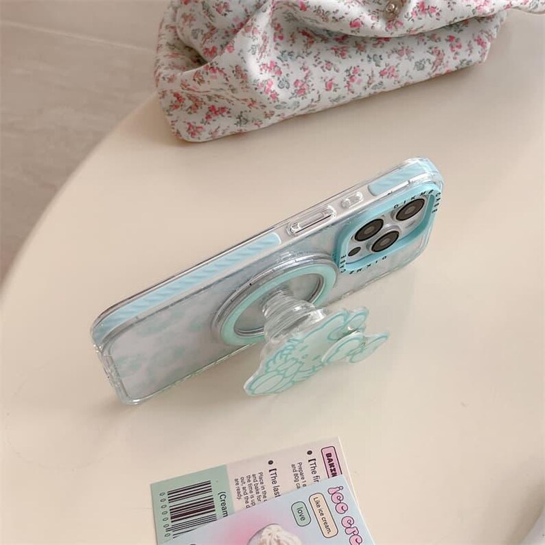 K&ouml;nnte beinhalten: Eine transparente Handyh&uuml;lle mit hellblauem Wolkenmuster und einem passenden Pop-up-Griff. Die H&uuml;lle hat einen hellblauen Rand und eine transparente R&uuml;ckseite. Der Pop-up-Griff hat die Form einer Wolke. Ein florales Gewebe befindet sich im Hintergrund.