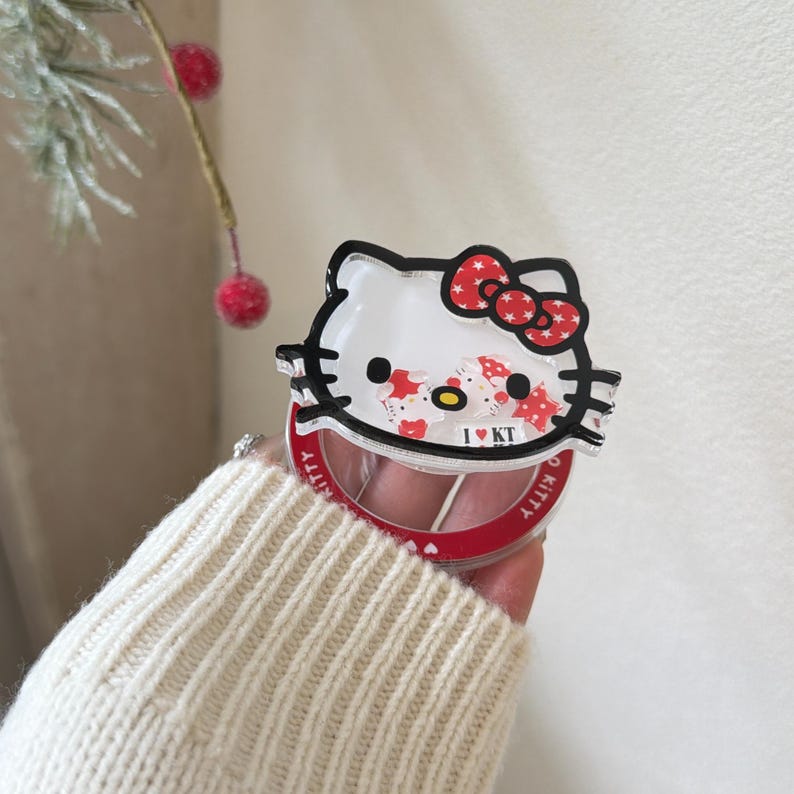 May include: A clear, red-rimmed compact mirror featuring a Hello Kitty design. The design includes a red bow, black whiskers, and the text "I KT". The mirror is held in a hand wearing a cream-colored sweater.
