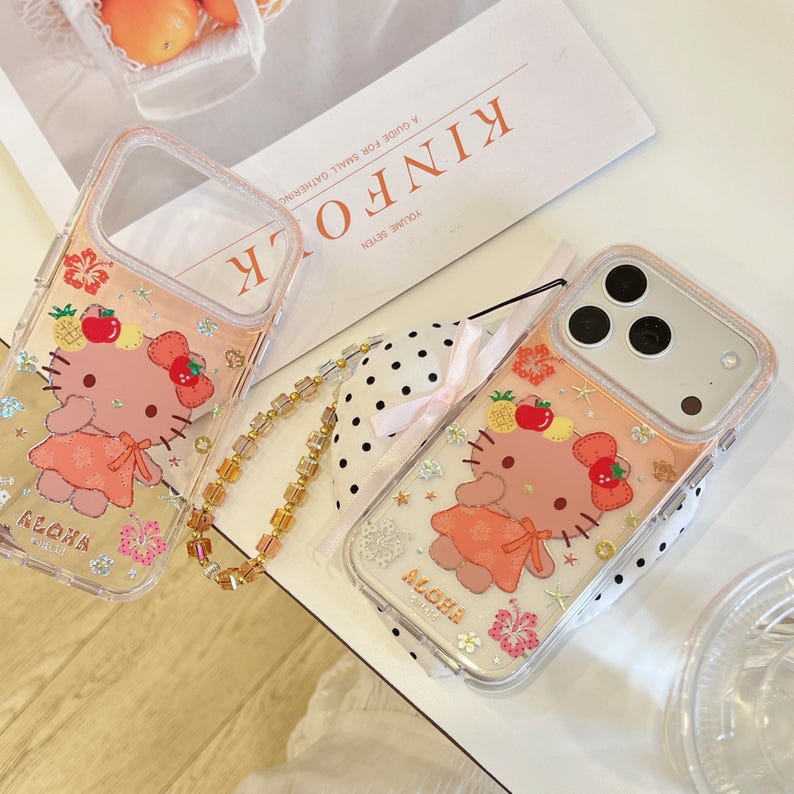 May include: Clear phone cases with a pink and orange design featuring a cartoon character, fruit, and floral accents. The cases have a gold-colored wrist strap and the word "ALOHA" is printed on the bottom.