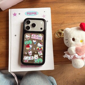May include: A black phone case with a clear back featuring a colorful Hello Kitty design. The case is on a white book with the word "Hello" printed on it. A Hello Kitty plush toy and gold keychains are also visible.